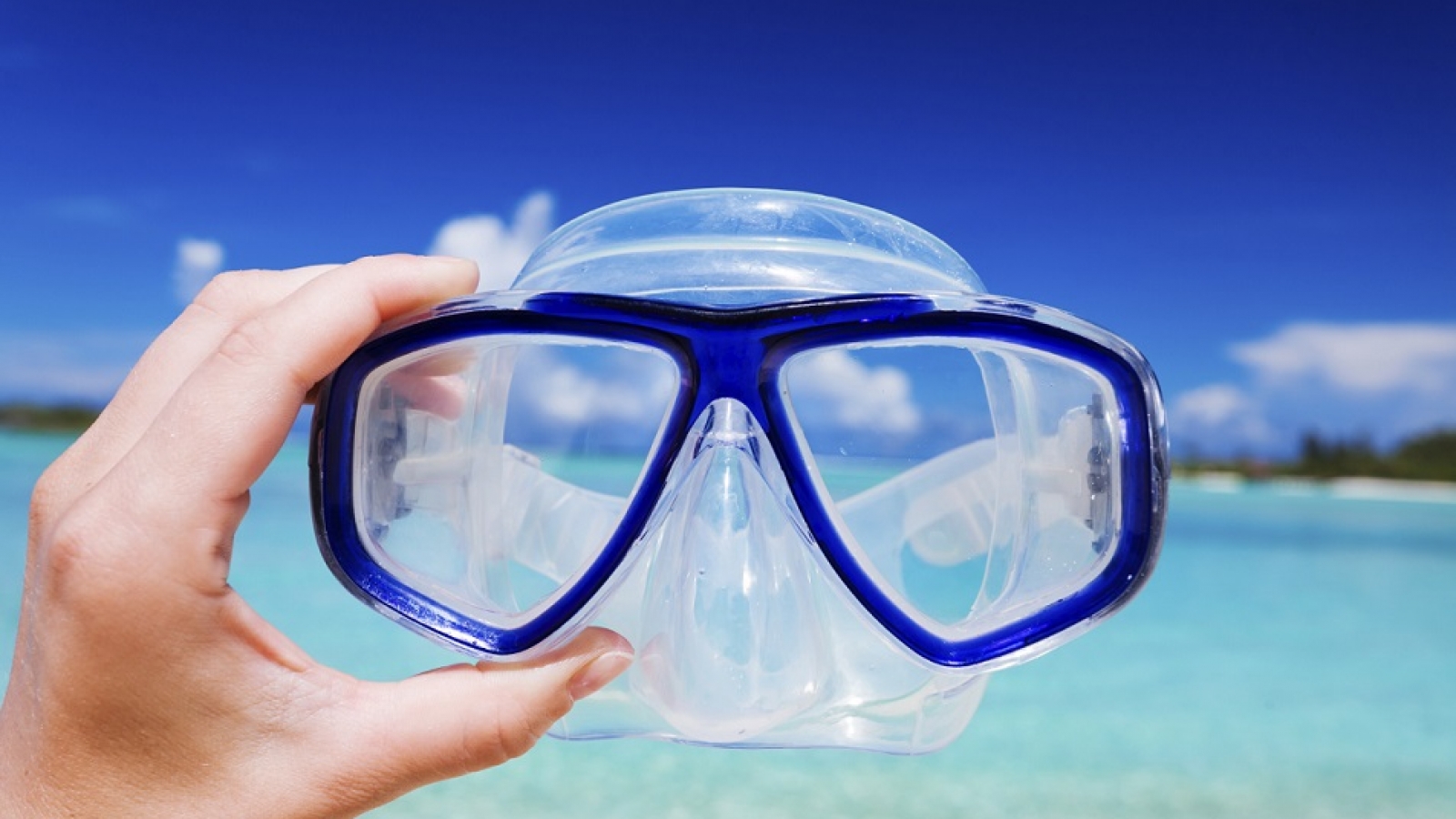 Hand holding snorkel googles against beach and sky